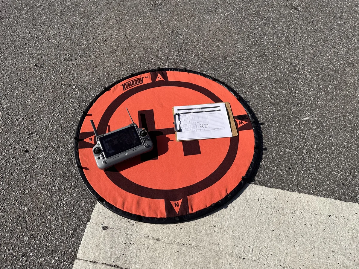 Top-down view of a Hoodman landing pad with the aircraft controller and clipboard holding the printed field log at the launch site for a multi-zone aerial mapping mission