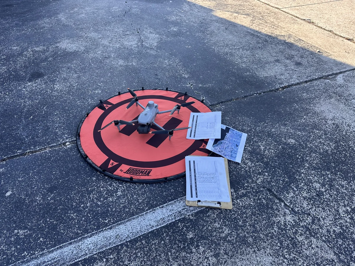 A field aircraft staged on a Hoodman landing pad with printed field logs, flight documentation, and an aerial map print staged on the adjacent pavement at the launch site