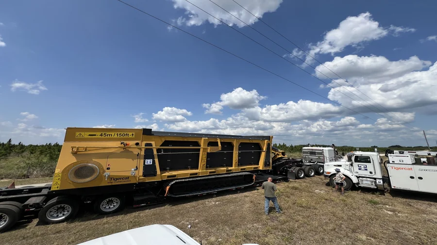 A yellow Tigercat 6040 Carbonizer mobile biochar production unit sits on a lowboy transport trailer in an open grassy field, accompanied by a white Tigercat T-143 hauler truck and a Tigercat RotoBec grapple saw in the background under a partly cloudy sky