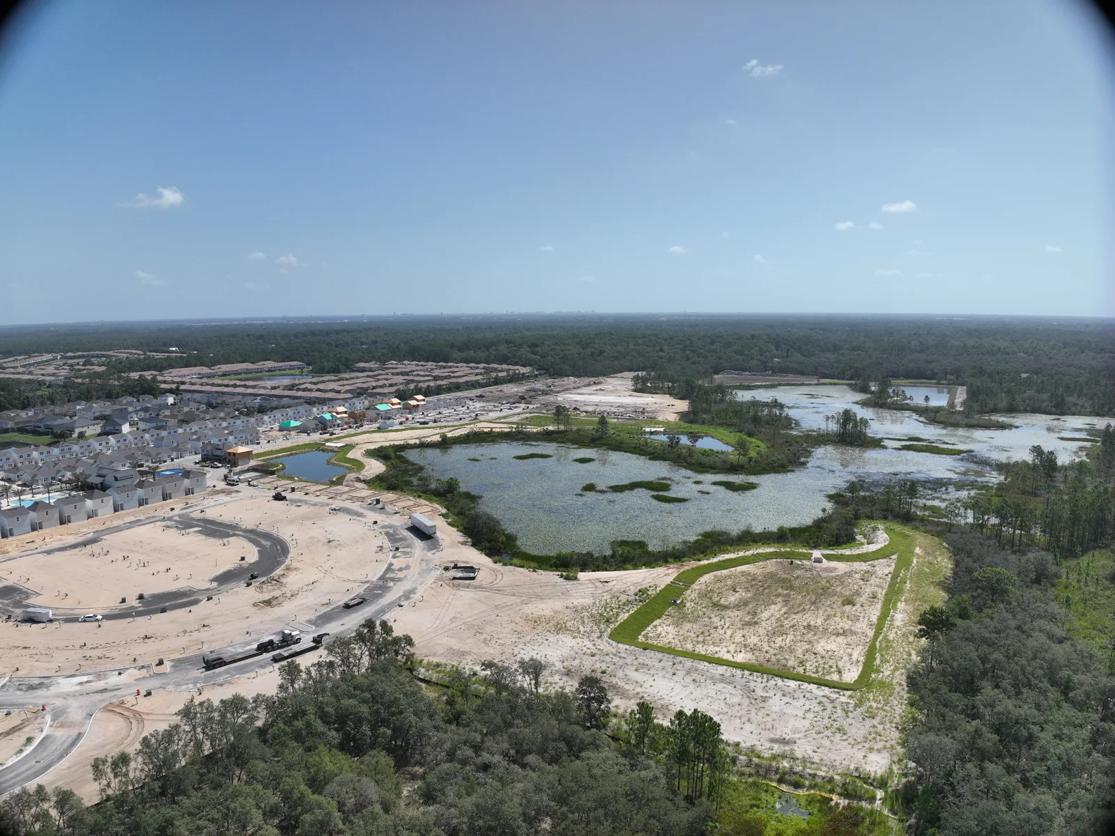 Wide aerial photograph of a master-planned residential community in mid-phase construction, showing completed single-family homes alongside active grading, new road construction, retention ponds, and preserved wetland areas in Central Florida.