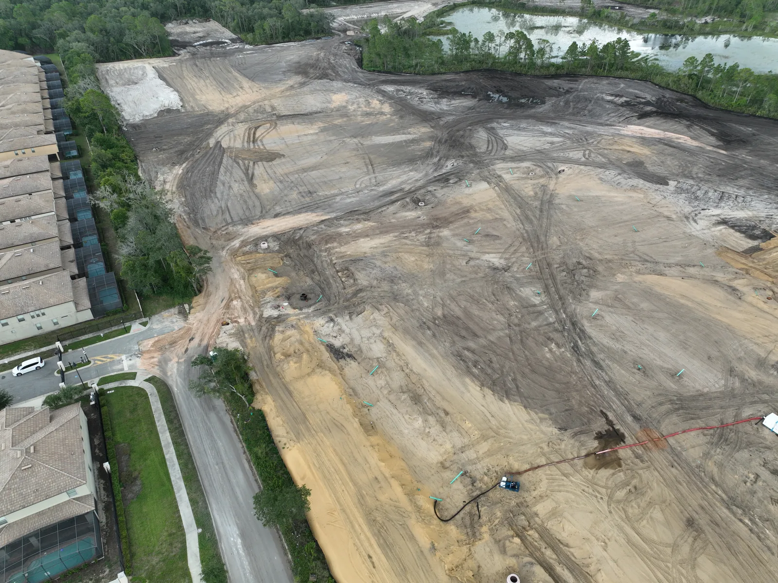 Oblique aerial photograph of an active residential development site in the mass earthwork phase, with exposed sandy and dark soil areas, sweeping equipment tracks across the disturbed ground, a retention pond in the background, and existing finished single-family homes visible on the left edge of the frame.